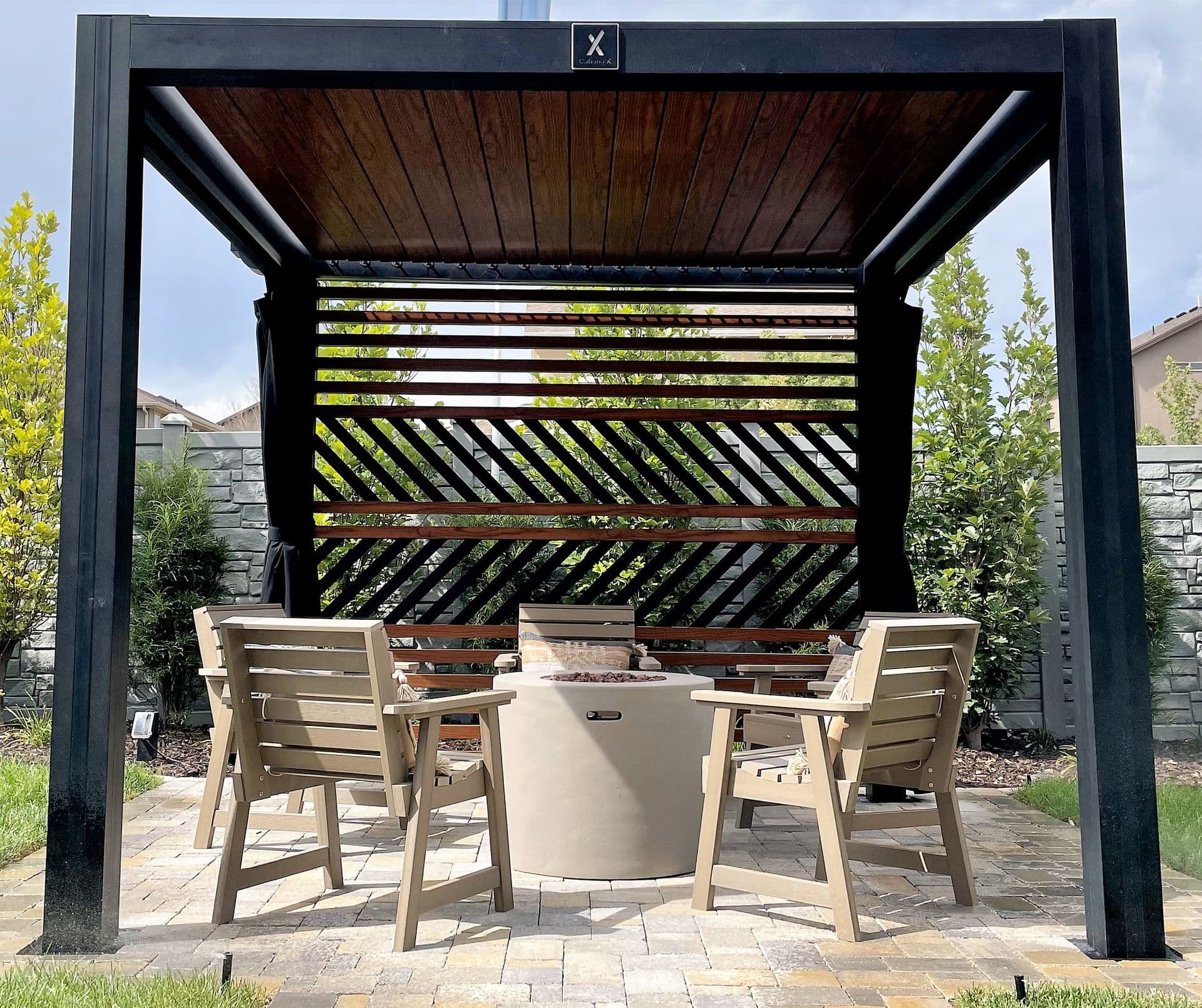A modern outdoor pergola with a wood slat roof shelters four wooden chairs around a round fire pit. Stone walls and lush greenery surround the scene.