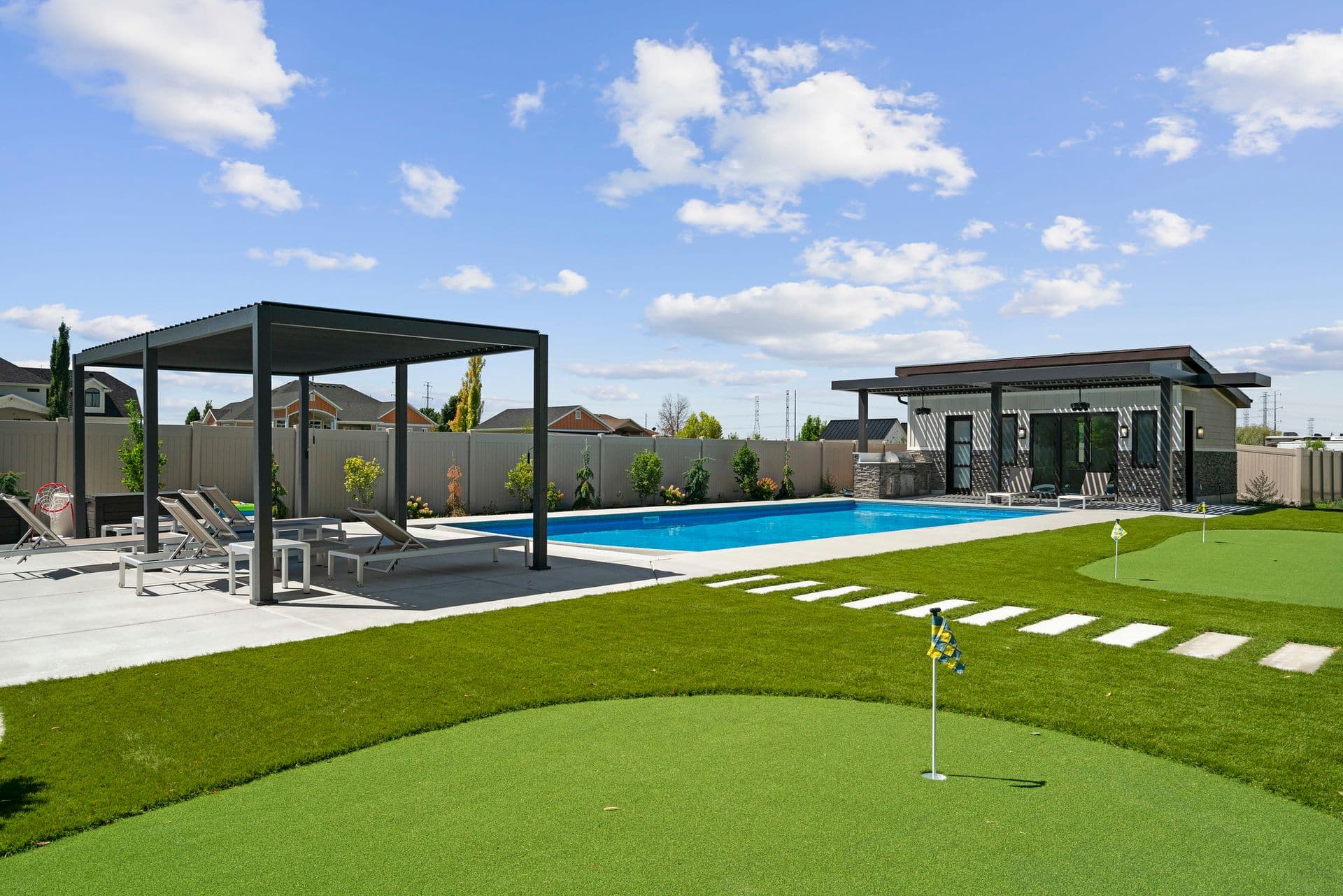Modern backyard with a rectangular pool, pool house, shaded lounge chairs, and a mini-golf putting green. Sunny day with scattered clouds.