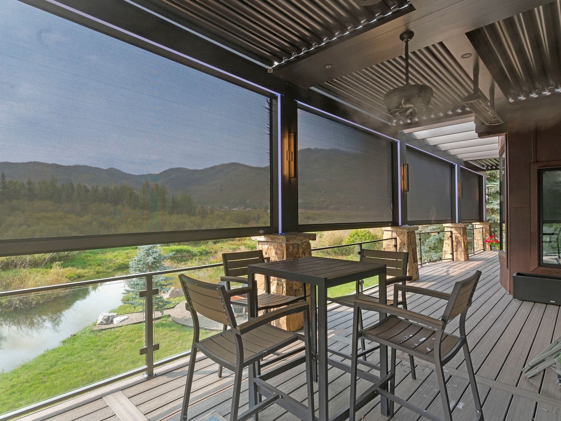 Stylish patio with high table and chairs, surrounded by mesh screens that reveal a scenic view of hills and a stream, creating a serene atmosphere.