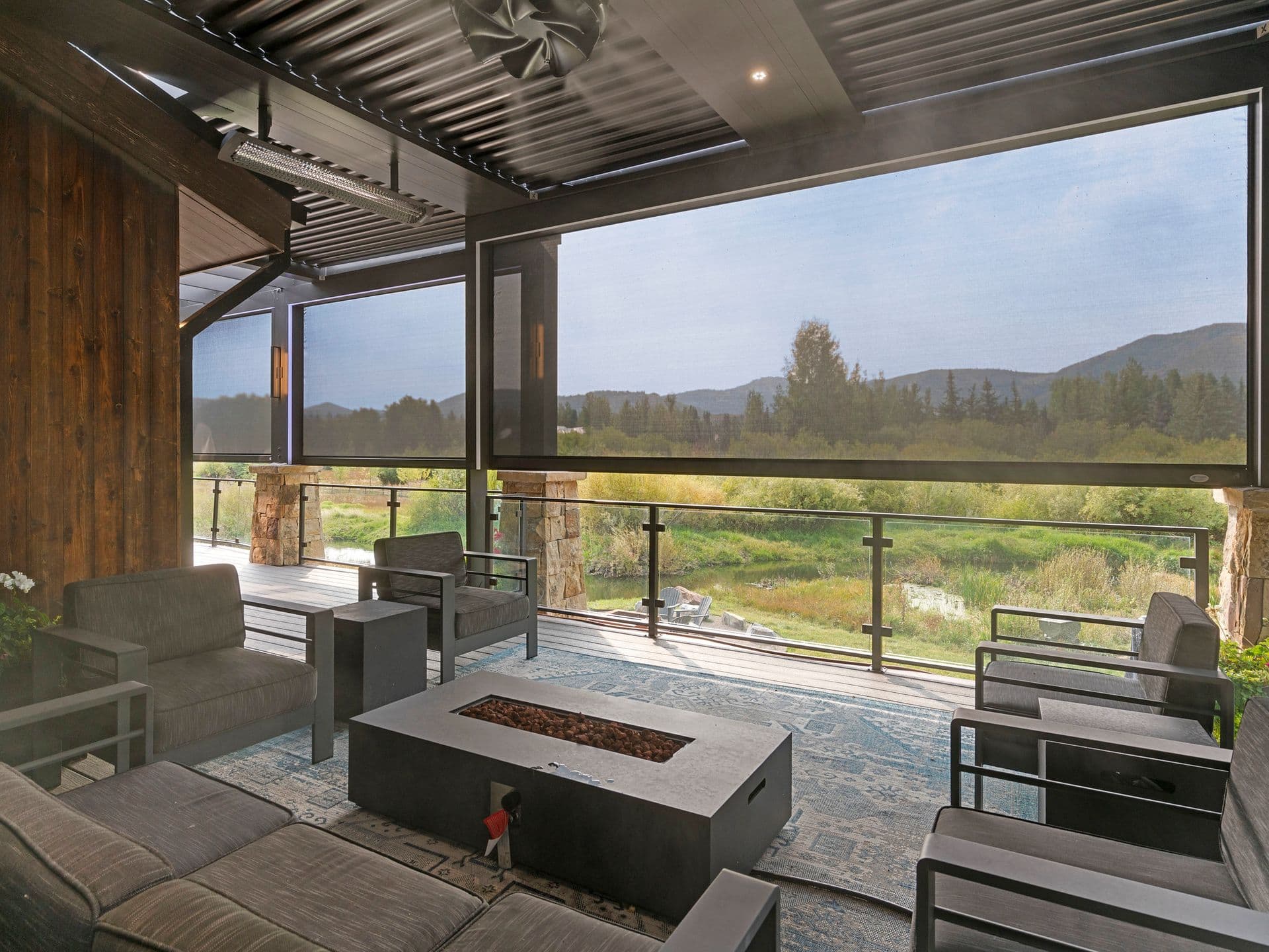 Modern outdoor patio with gray sofas and a rectangular fire pit. Overlooks a scenic view of lush greenery and distant mountains through a glass railing.