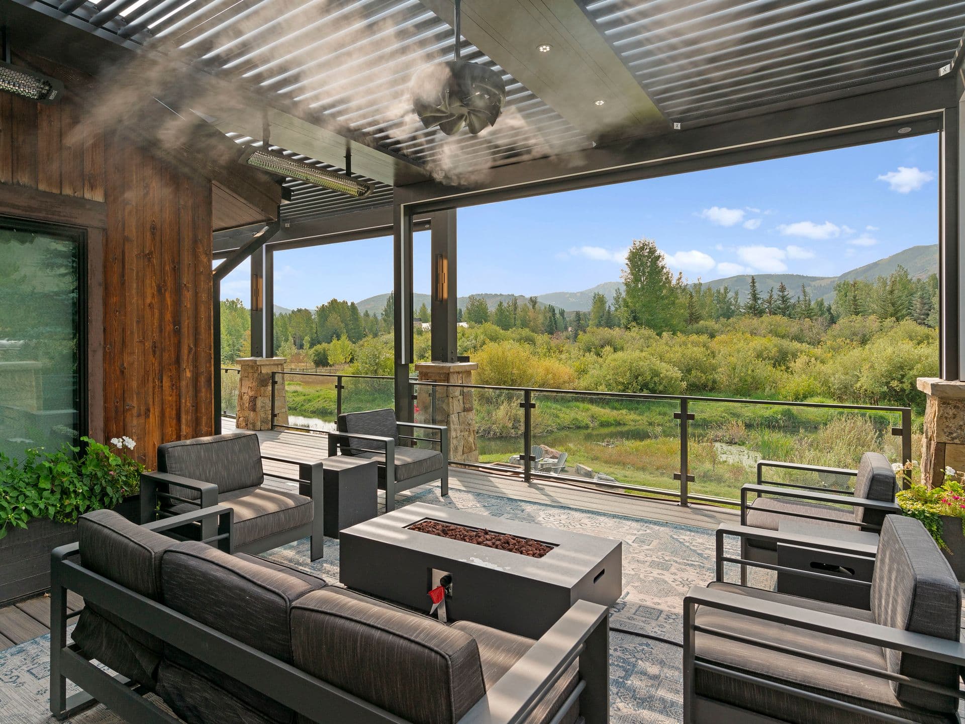 Modern covered patio with sleek black furniture, a fire table, and misting fan. Overlooks lush green landscape and distant mountains under a clear blue sky.
