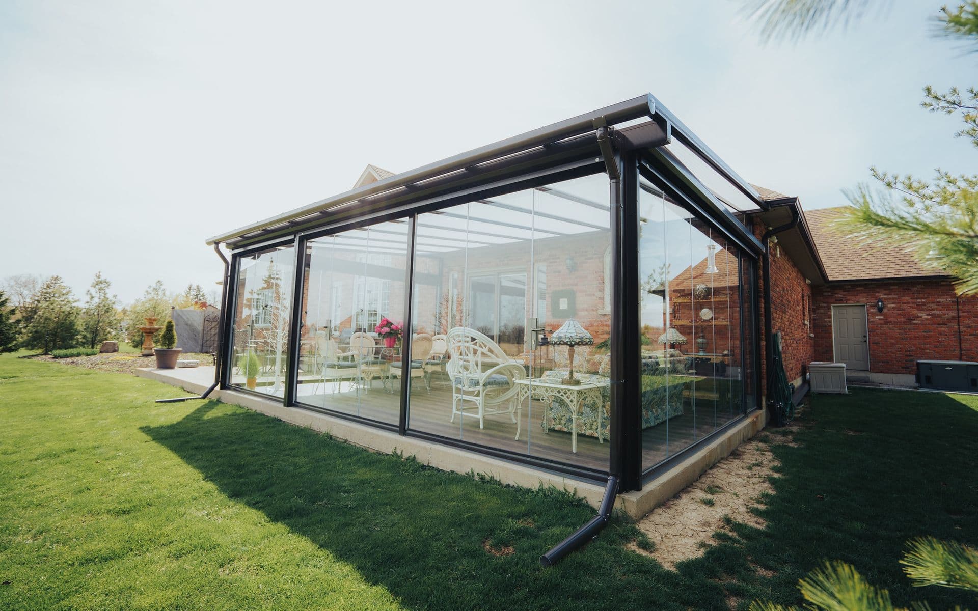 A modern sunroom with glass walls attached to a brick house, showcasing white wicker furniture inside. The surrounding garden is lush and green, conveying tranquility.