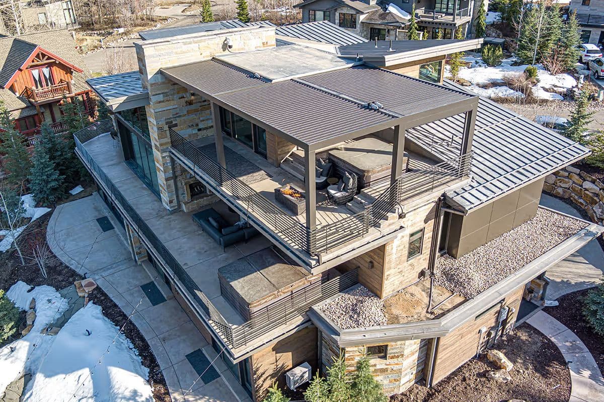 Aerial view of a modern, multi-story house with stone and metal exterior. It features large terraces, surrounded by trees and snowy ground. Winter setting.