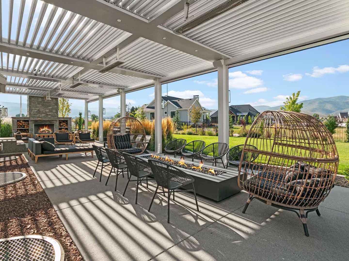 Modern outdoor patio with a pergola, wicker chairs, and a long fire pit. A stone fireplace and green garden create a cozy, inviting atmosphere.