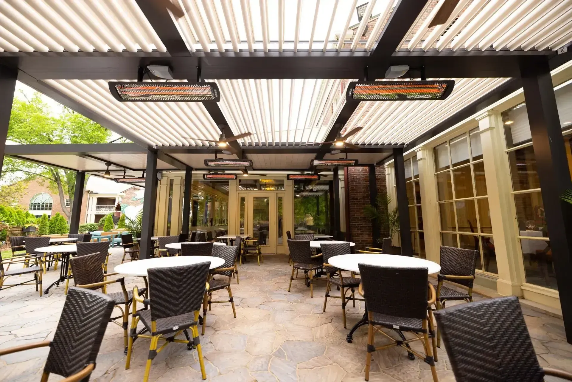 Outdoor patio with stone flooring, covered by a pergola with heaters. Empty white round tables surrounded by wicker chairs create a cozy ambiance.